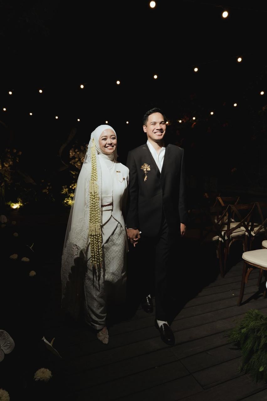 A bride in a hijab and ivory kebaya holding a strand of melati, gold brooches at her collar.