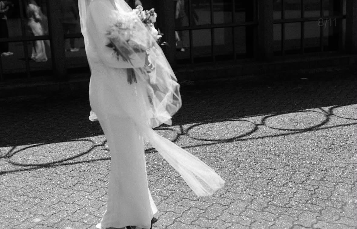 A bride walking in a narrow white dress with a light scarf moving behind her.